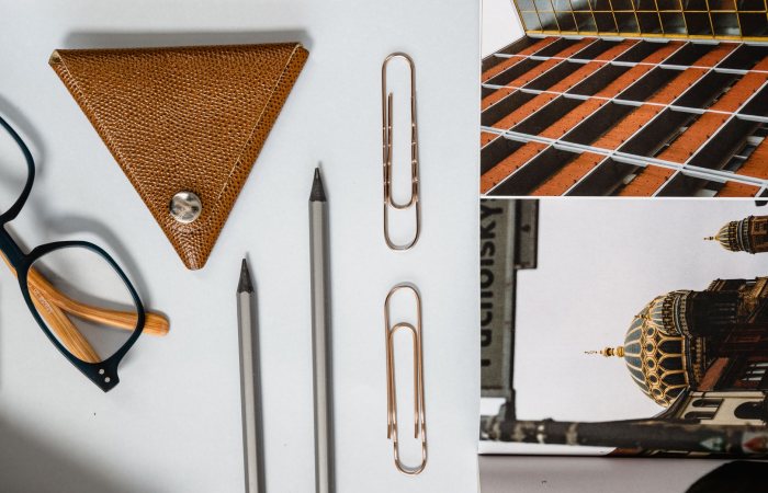 Flat lay of eyeglasses, pencils, paper clips, and leather keyholder on a white background with architectural postcard.