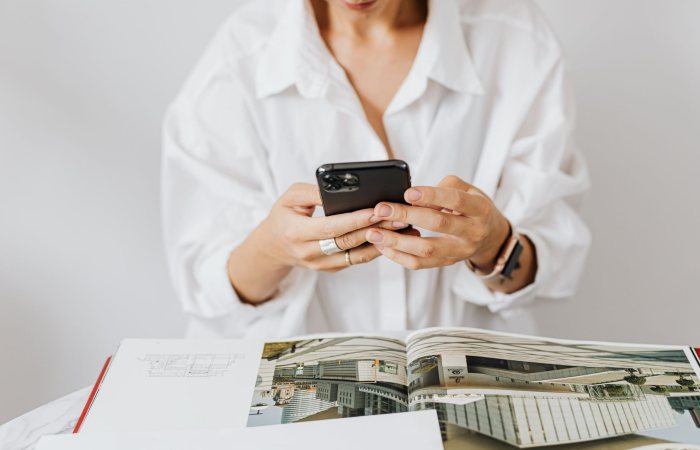 Woman in white shirt using smartphone while reading architecture magazine. Indoor setting.